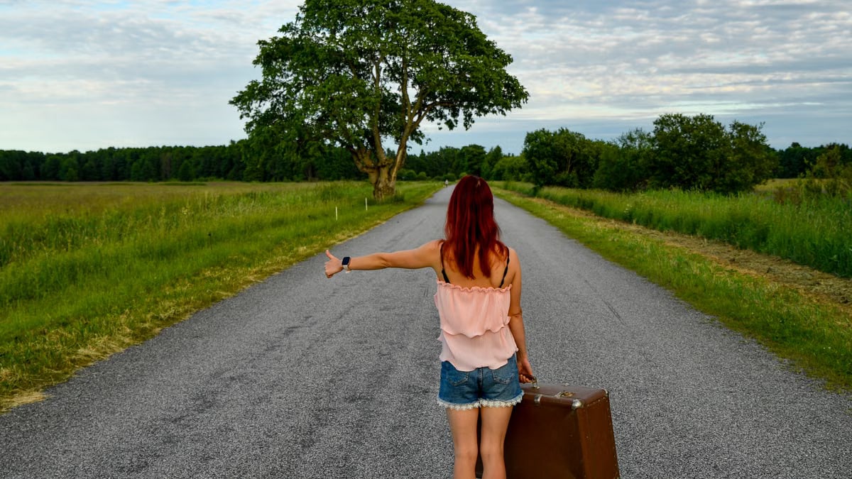 A woman with red hair hitchhiking on a country road in Estonia with a vintage suitcase.